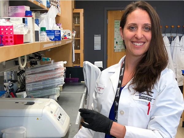 Researcher in a lab coat smiling while holding a sample plate in a laboratory.