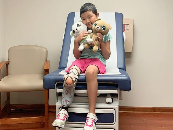 Young child with a prosthetic leg sitting on an exam table, holding two stuffed animals and smiling.