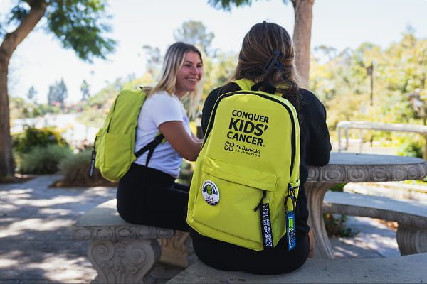 Two people sitting outdoors, wearing bright green backpacks that reads “Conquer Kids’ Cancer.”