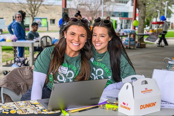 Two smiling volunteers in St. Baldrick’s shirts sit at a table with a laptop at an outdoor event.