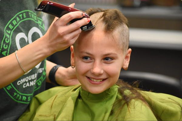 A smiling child having their head shaved with electric clippers while wearing a green barber cape.