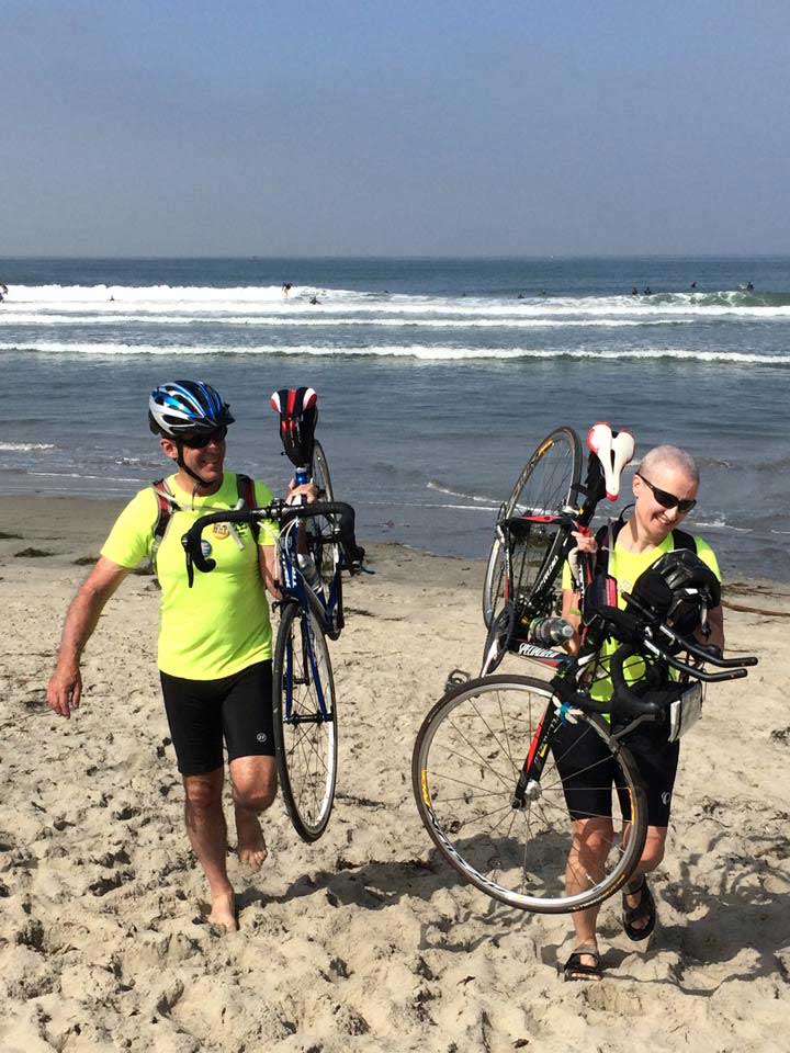 Kim and Jon lift their bikes on their shoulders at the Pacific Ocean at the start of their ride